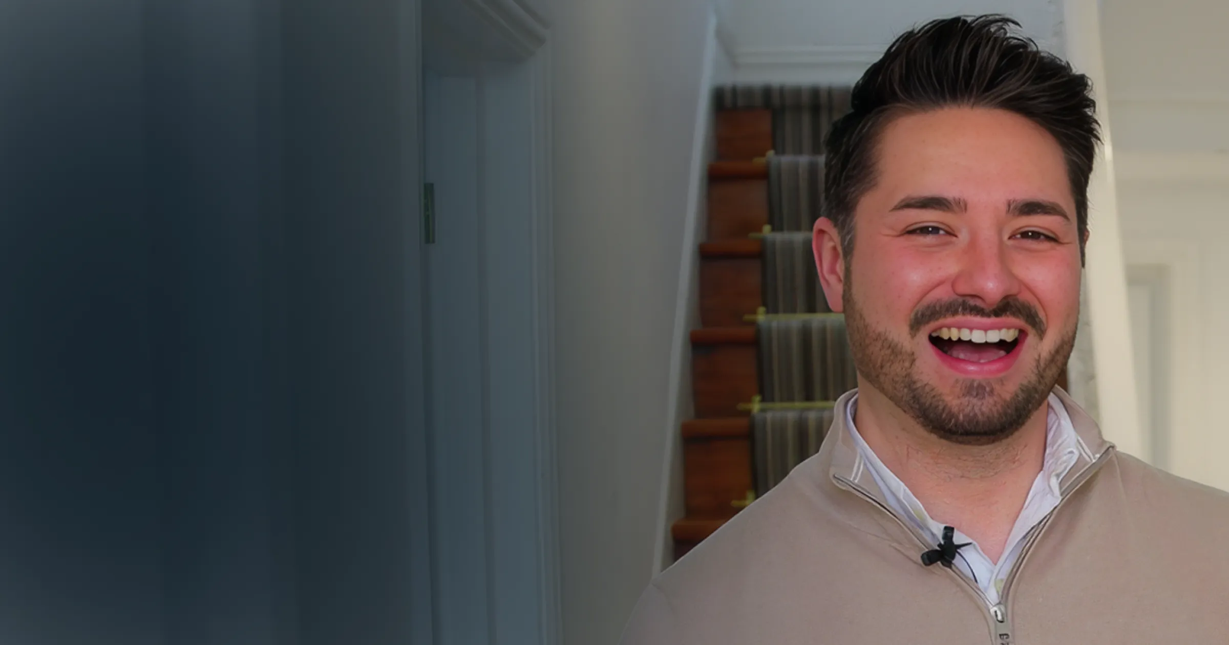 A man with short dark hair and a beard smiles warmly, wearing a light zip-up jacket. A wooden staircase with a patterned runner is in the background.
